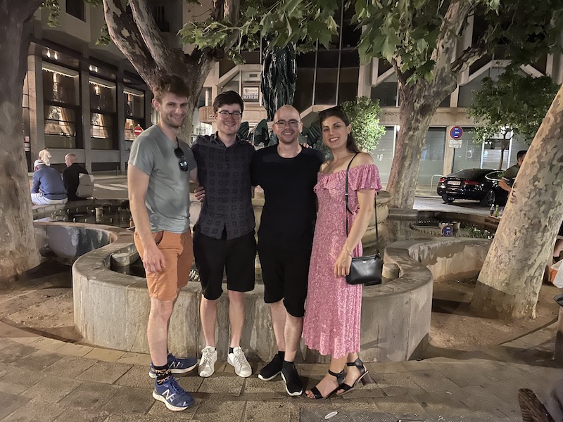 four people stand wearing summer clothes smiling at the camera in front of trees in a town square
