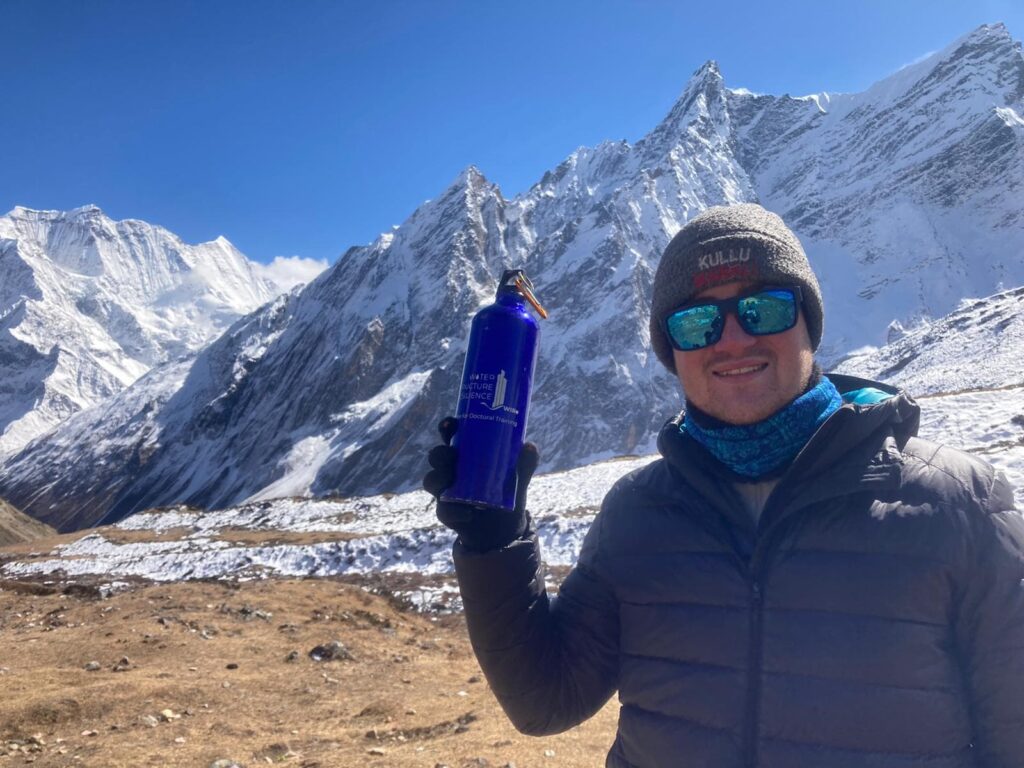 Matthew stands smiling at the camera wearing a blue coat, a hat and blue sunglasses. in the background there are snowy mountains. Matthew is holding a water bottle with the WIRe logo