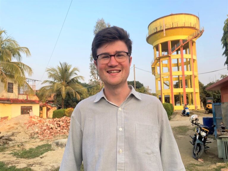 Matthew stands in the centre of the frame wearing a pale blue shirt and glasses smiling at the camera in the background there are palm trees