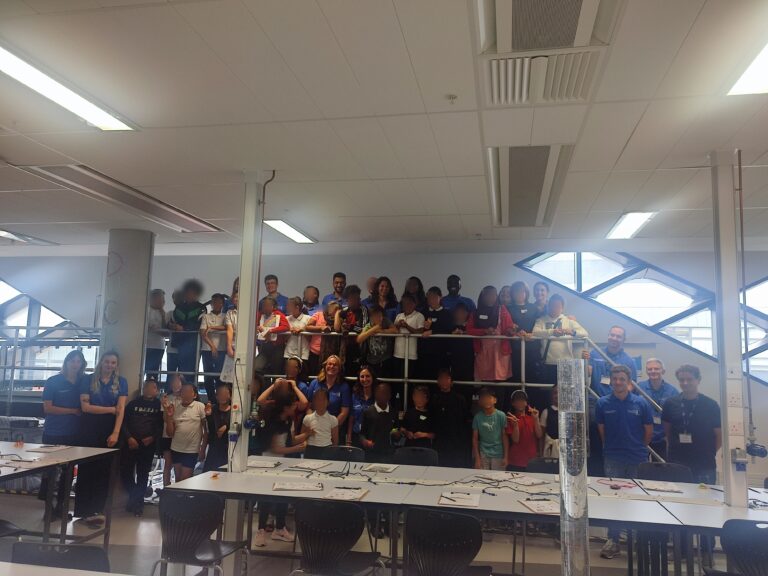 a group of children and PhD students stand in a science classroom smiling at the camera