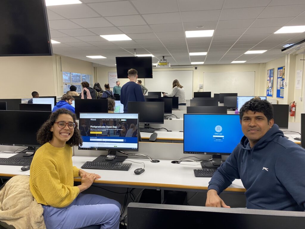 Isabel and Ishara sit in a room of computers on desks, they are sat in front of two computers smiling at the camera. Isabel wears a yellow top and Ishara wears a grey hoodie