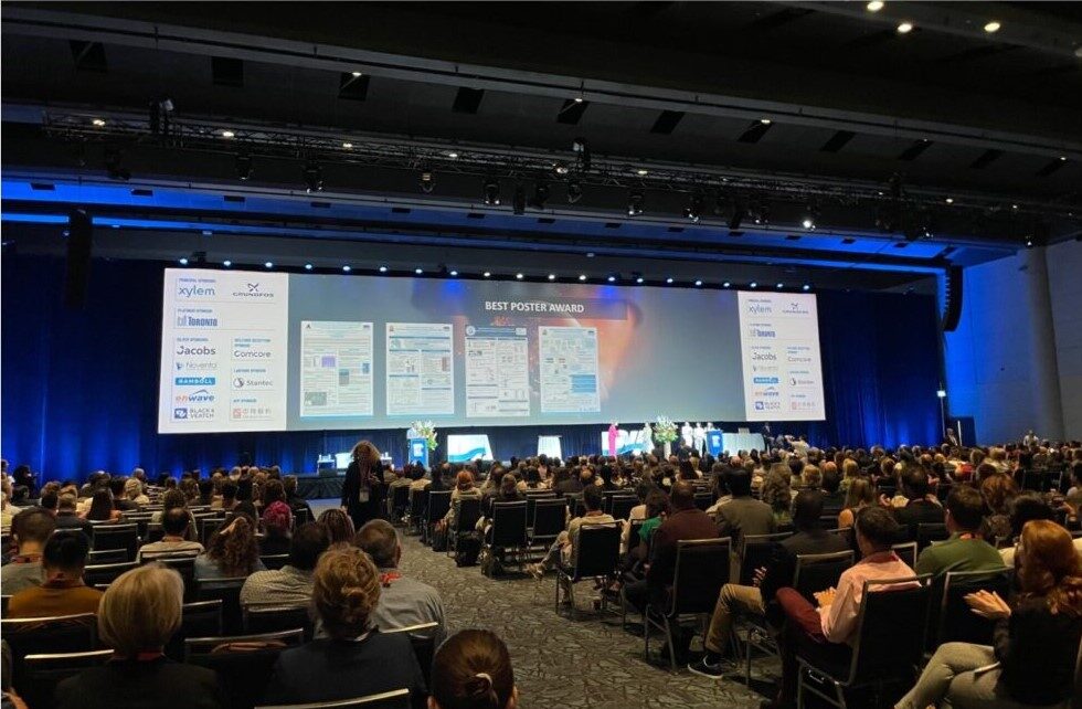 A large room of people in chairs facing a big screen announcing best poster awards