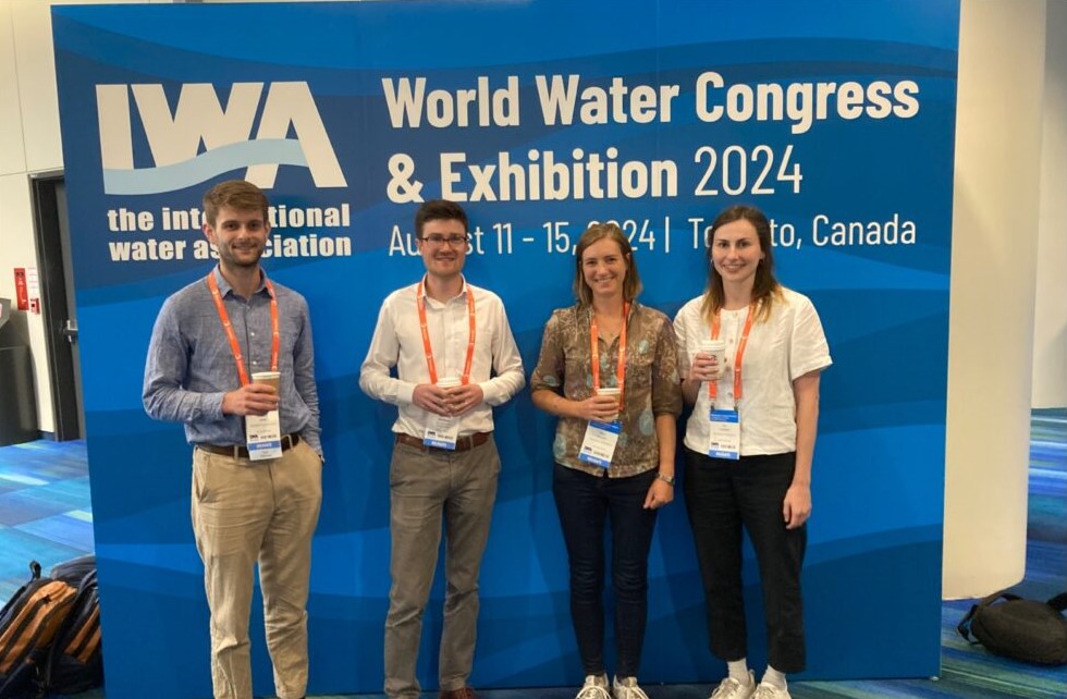 Edward, Matthew, Polly and Ali stand smiling at the camera in front of an IWA World Water Congress and Exhibition 2024 banner