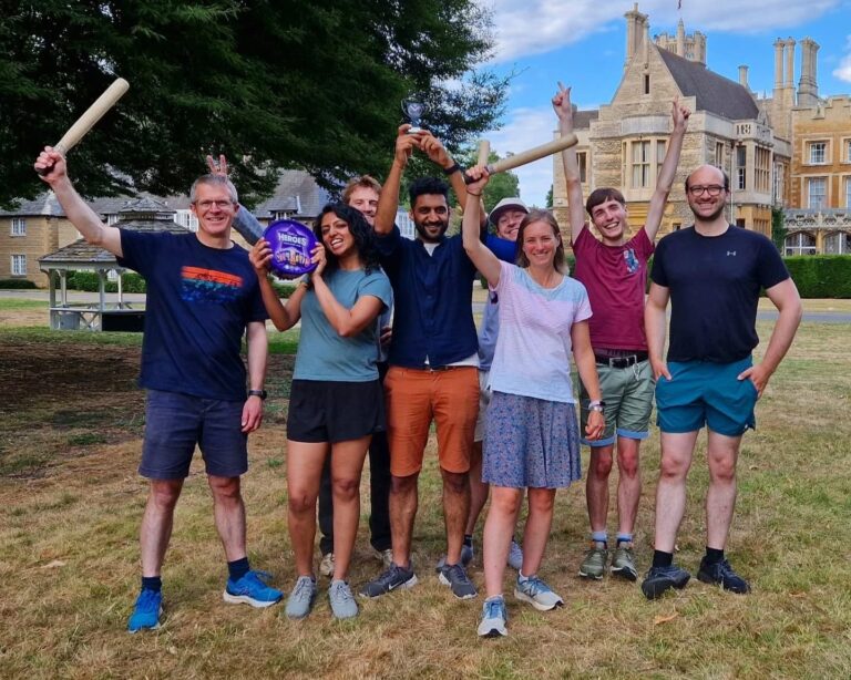A group of people stand smiling at the camera outside in the sun. some are holding rounders bats. one is holding a trophy and another is holding a box of chocolates