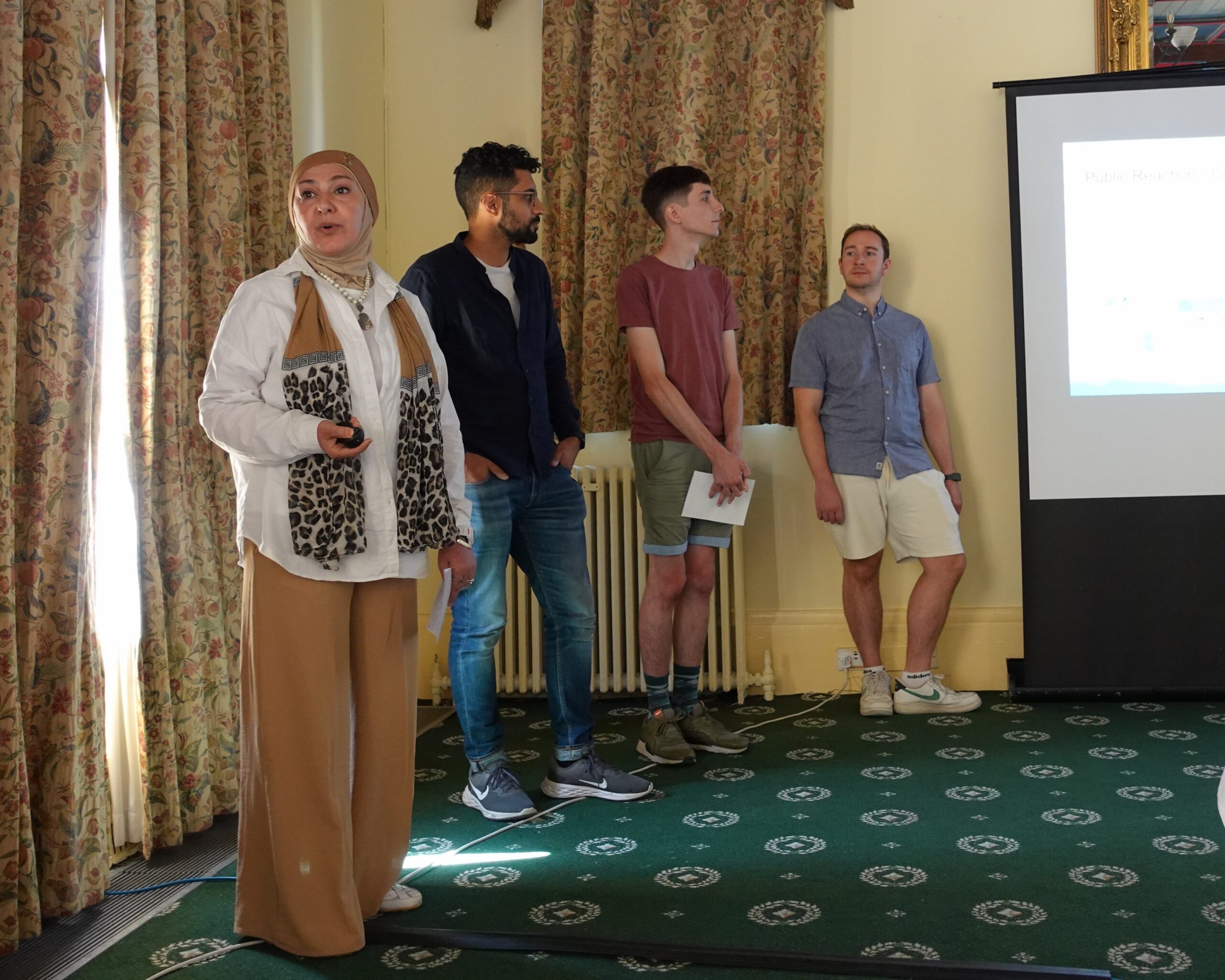 Group 4 presenting their summer challenge proposal. on the left of the field of view a woman wearing a white shirt, brown trousers, and a brown and leopard print headscarf talks to the audience (off camera). three people stand behind her