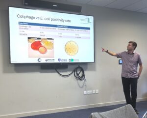 A man presents, he is pointing at a screen with a powerpoint slide. the audience is behind the photographer and out of shot