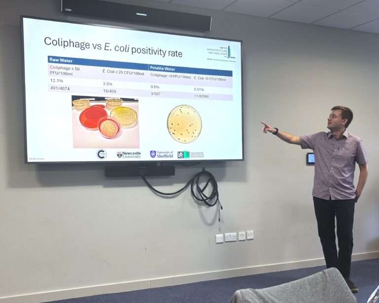 A man presents, he is pointing at a screen with a powerpoint slide. the audience is behind the photographer and out of shot