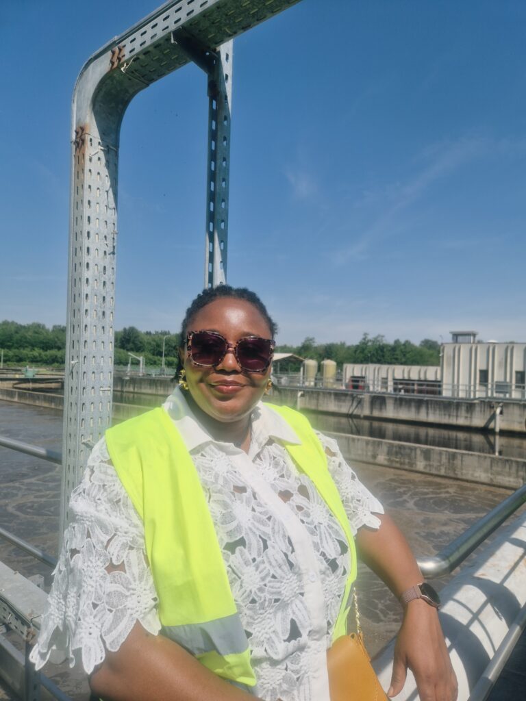 a woman smiles and poses for a picture in front of a water treatment facility, she is wearing a white top and a yellow high visibility vest
