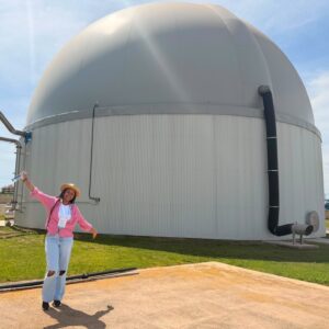 a woman stands smiling in front of a large domed building at an industrial site. the sun is shining. the woman is holding her arms out wide.