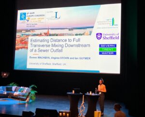 A woman stands on a stage in front a large screen. she is stood at a lectern delivering a talk. the audience is not pictured.