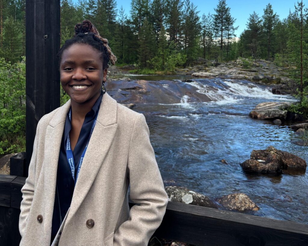 a woman stands smiling at the camera. Behind her is a river with trees on the banks. the sky is blue.