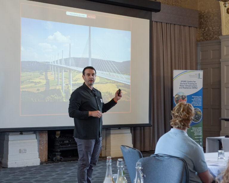 A man stood in front of a projector screen presents to an audience that is not in frame.