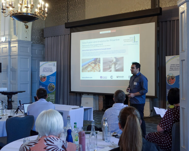 a man stands in front of a projector screen presenting to a room of people who are sat around round tables