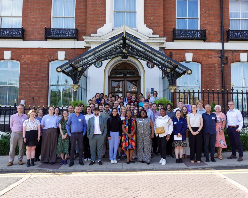 A large group of people wearing business attire stand in rows smiling at the camera. They are outside on grass, the weather is sunny with blue skies and behind them is a doorway to a red brick building. They are delegates from the CDT WIRe Annual One-Day Conference