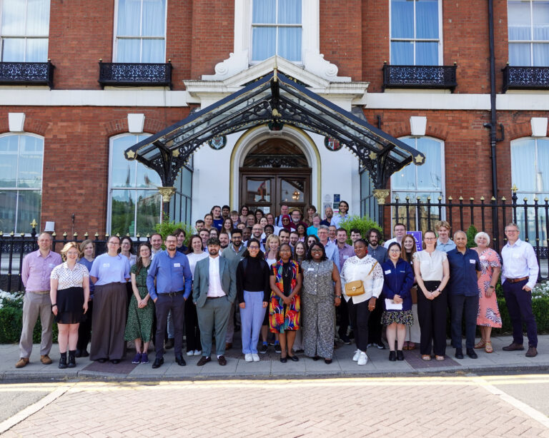 A large group of people wearing business attire stand in rows smiling at the camera. They are outside on grass, the weather is sunny with blue skies and behind them is a doorway to a red brick building. They are delegates from the CDT WIRe Annual One-Day Conference