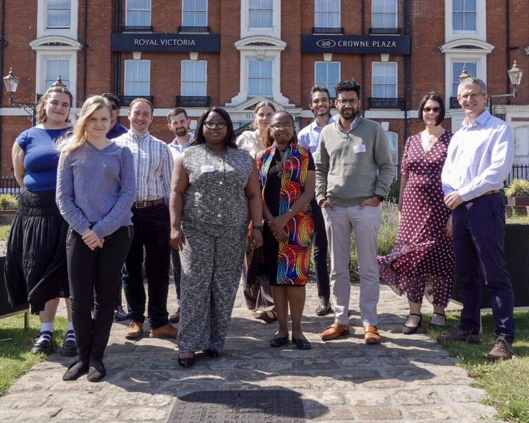 A group of people stand smiling at the camera, they are outside in the sun, stood in front of a red brick building.