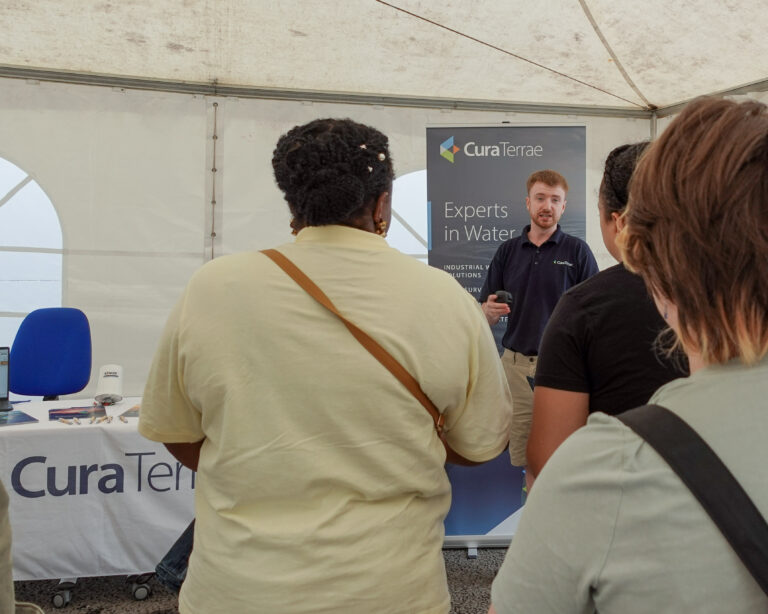A man talking to an audience. The audience has their backs to the camera. The man is stood at an exhibition stand that is branded with 'Cura Terrae'