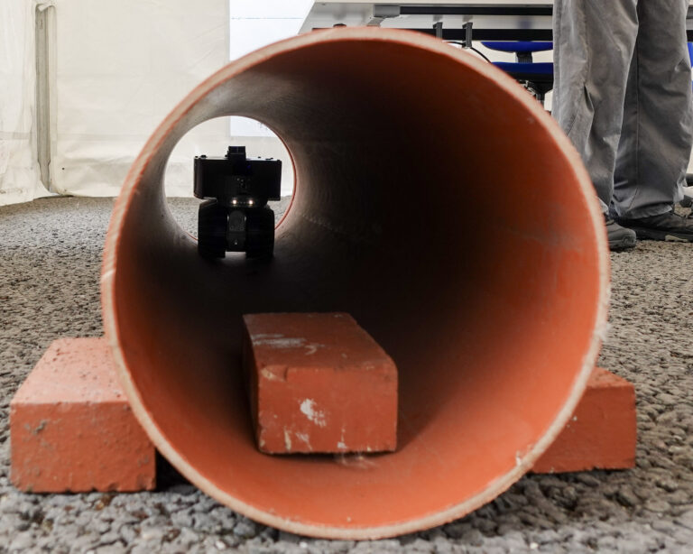 A small robot with wheels and lights on the front of it is in an orange plastic pipe