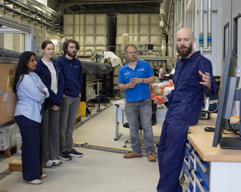 A man in a blue boiler suit talks to a group of people stood around a computer. They are in an industrial laboratory.