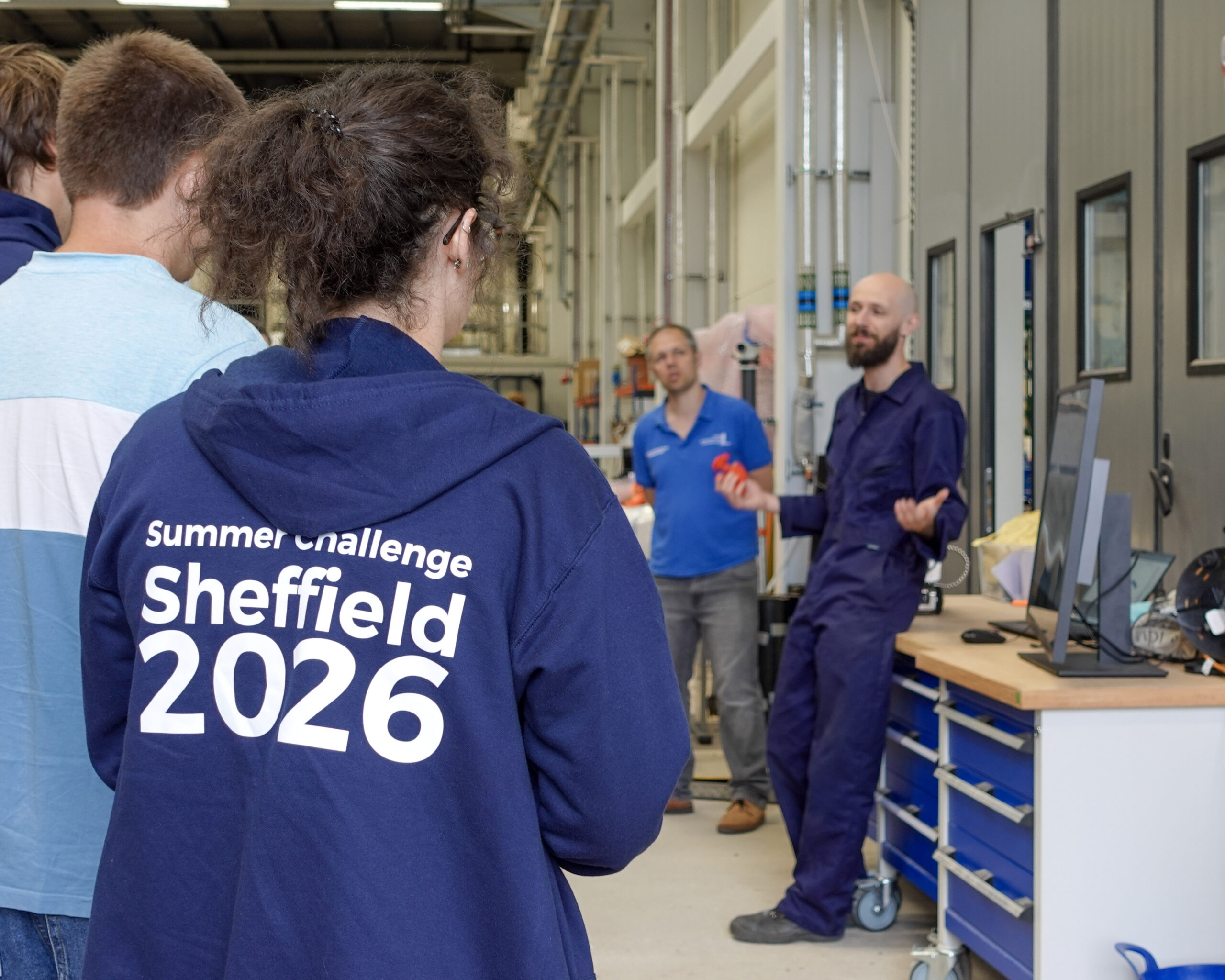A person is stood with their back to the camera listening to an industry demonstration. Text on the back of the hoodie in the foreground reads 'Summer Challenge Sheffield 2026'