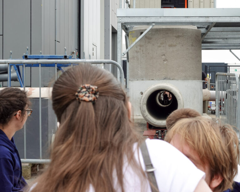 a group of people are looking down a concrete pipe to see a robot in the pipe. the picture is taken from behind the people