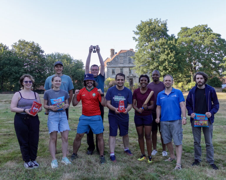 A group of people stood in a field smiling at the camera. One man lifts a trophy above his head