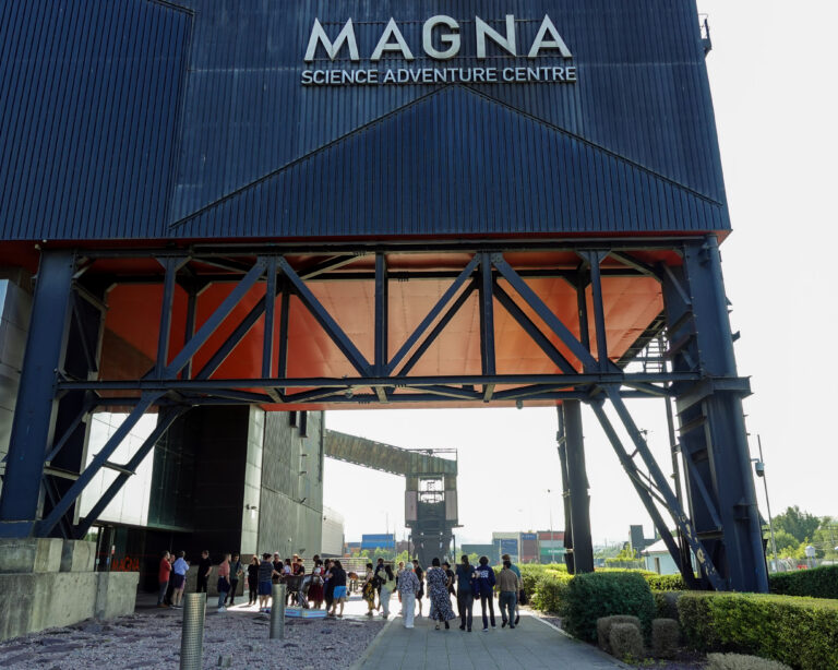 A large black building. A group of people walk below it. Text reads 'MAGNA SCIENCE ADVENTURE CENTRE'
