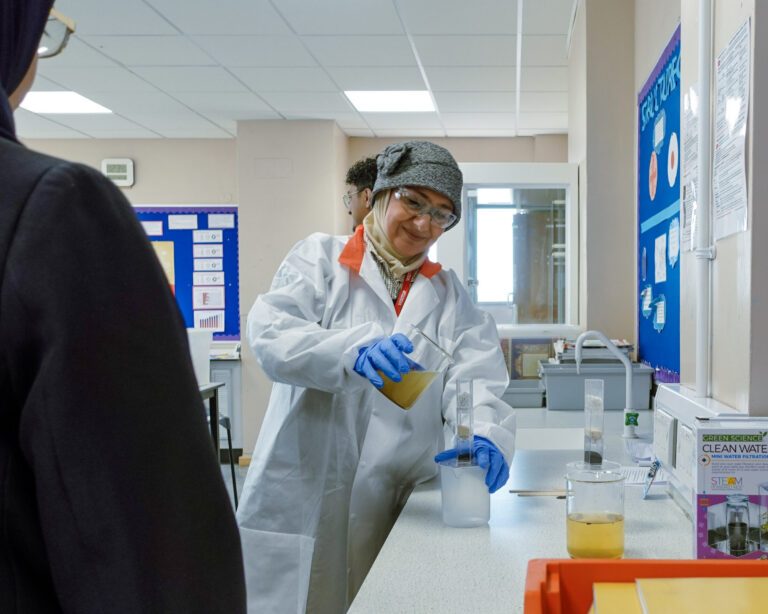 A woman wearing safety glasses, a lab coat, and blue gloves, pours a beaker of water during a science experiment in a classroom