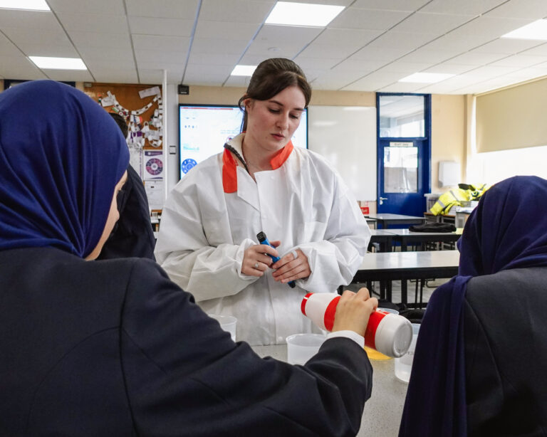 A woman stands behind two school students, who have their back to the camera. one student is pouring table salt in a science experiment