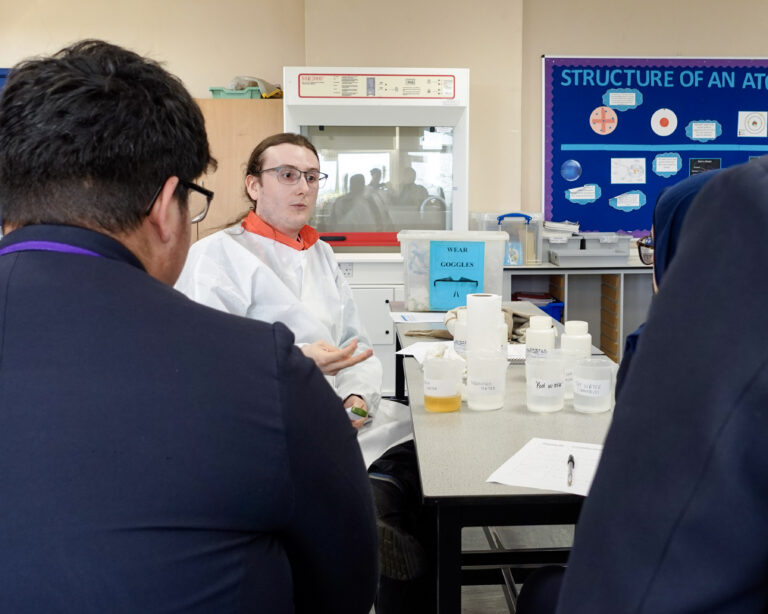A man sits in front of two school students who have their back to the camera. They are in a school science lab