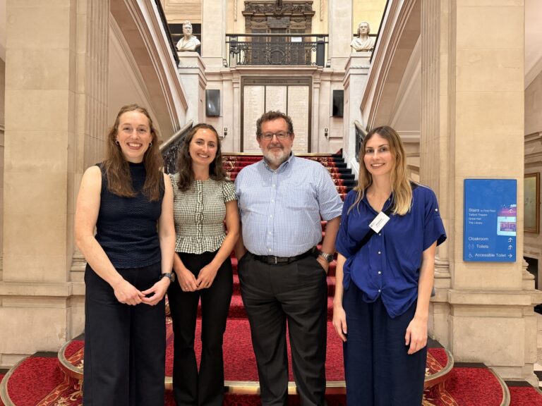 Four people stand smiling at the camera. They are at the bottom of a grand staircase.
