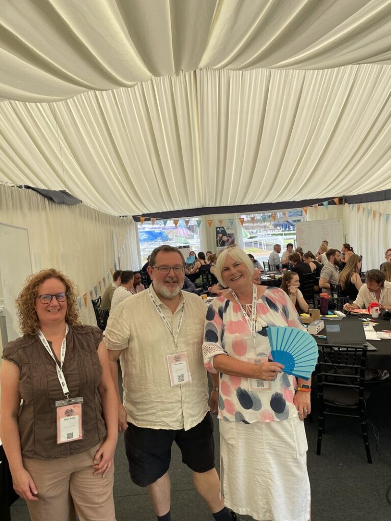 three people stand in a row smiling at the camera. They are in a marquee. The person on the right is holding a blue paper fan.