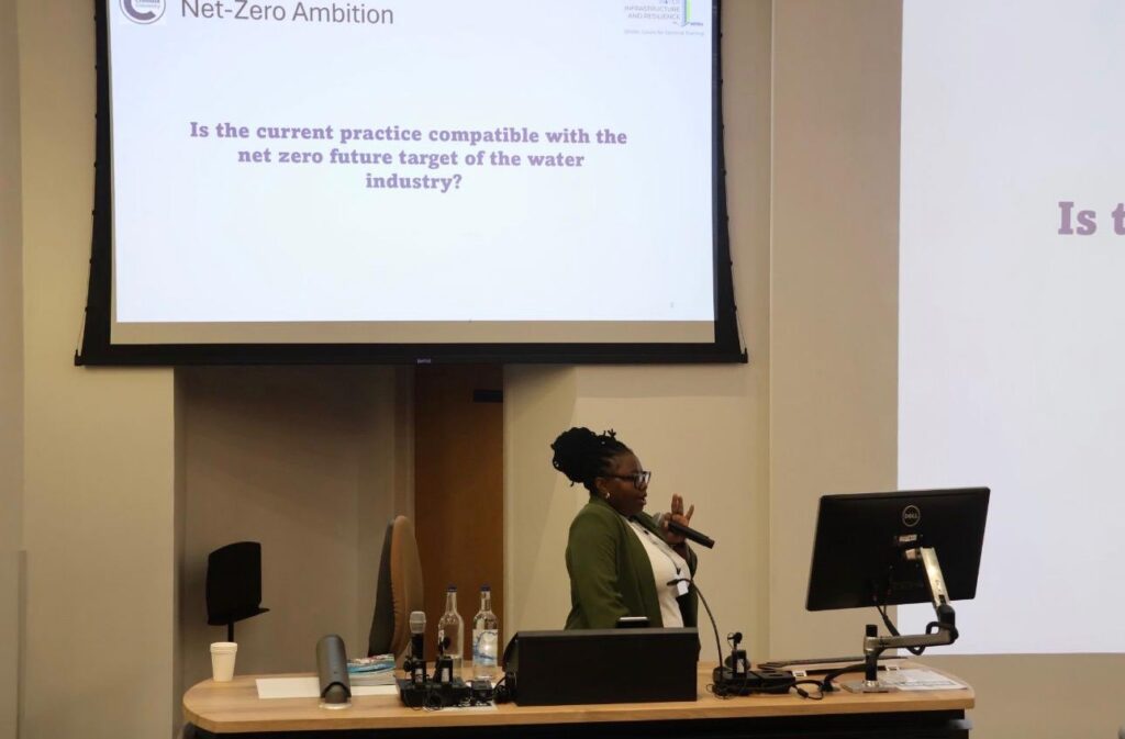 a woman stands in front of a projector screen. she is holding a microphone and presenting.