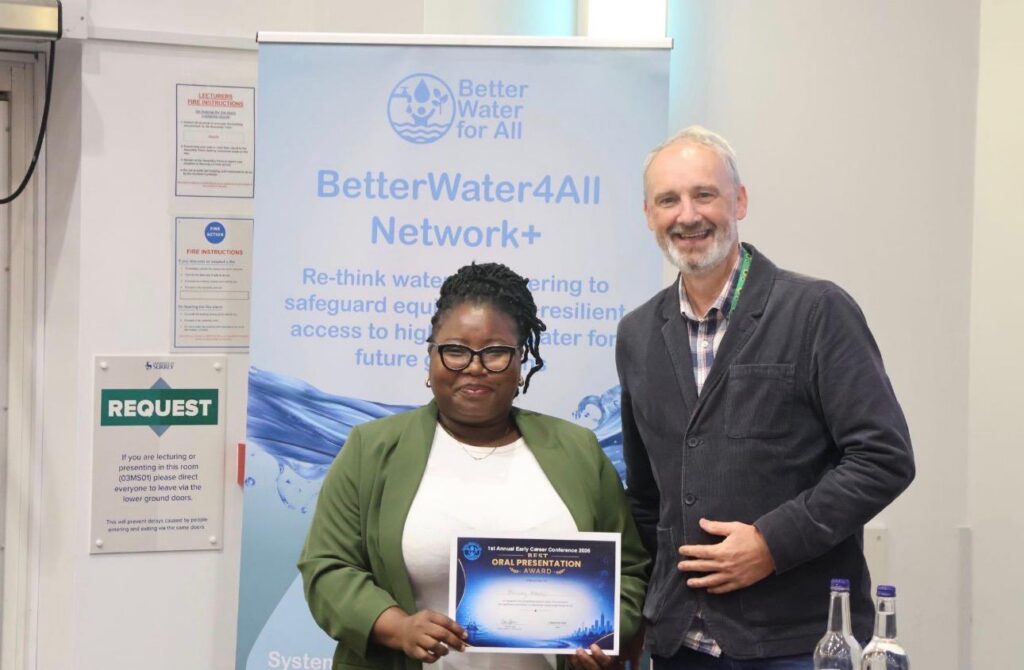 A man and a woman stand side-by-side smiling at the camera. the woman is holding a certificate.